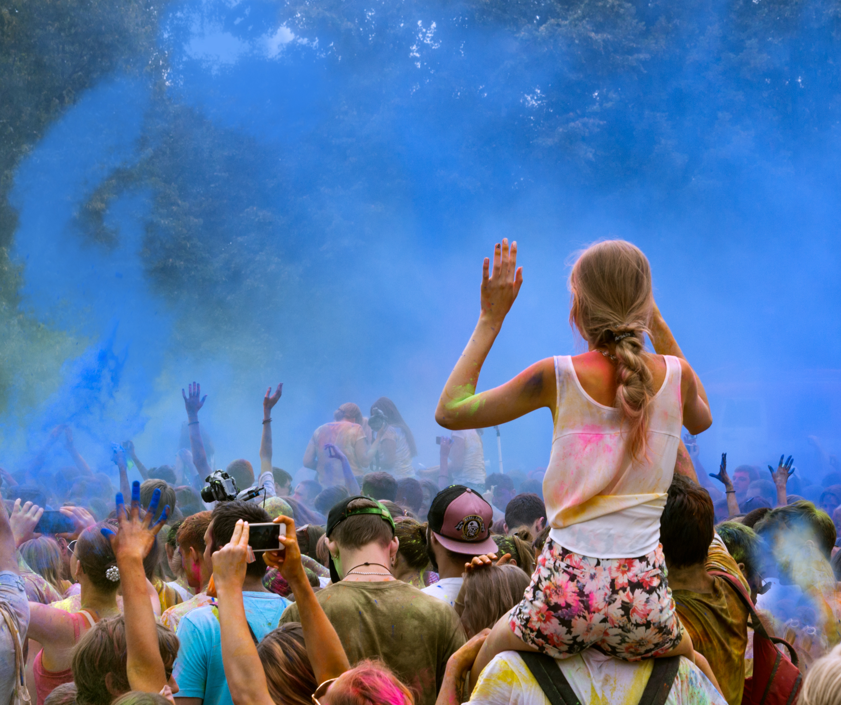 A crowd at a festival covered in color powder, with a girl on someone's shoulders raising her hands amid a burst of blue color.