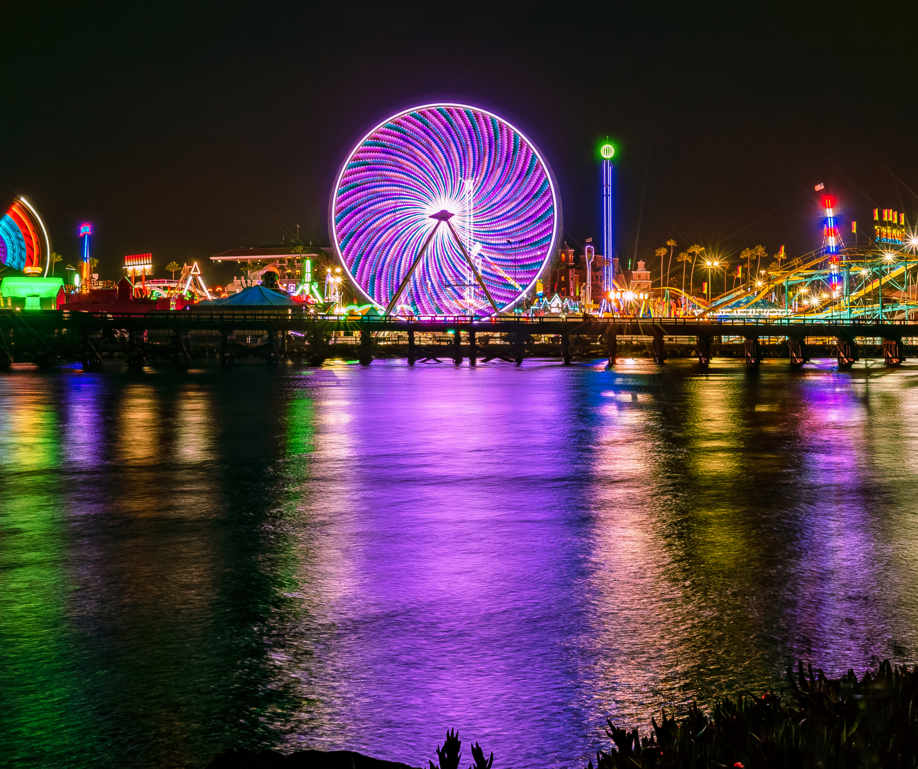 Nighttime view of a brightly lit Ferris wheel and amusement park with colorful lights reflecting on the water.