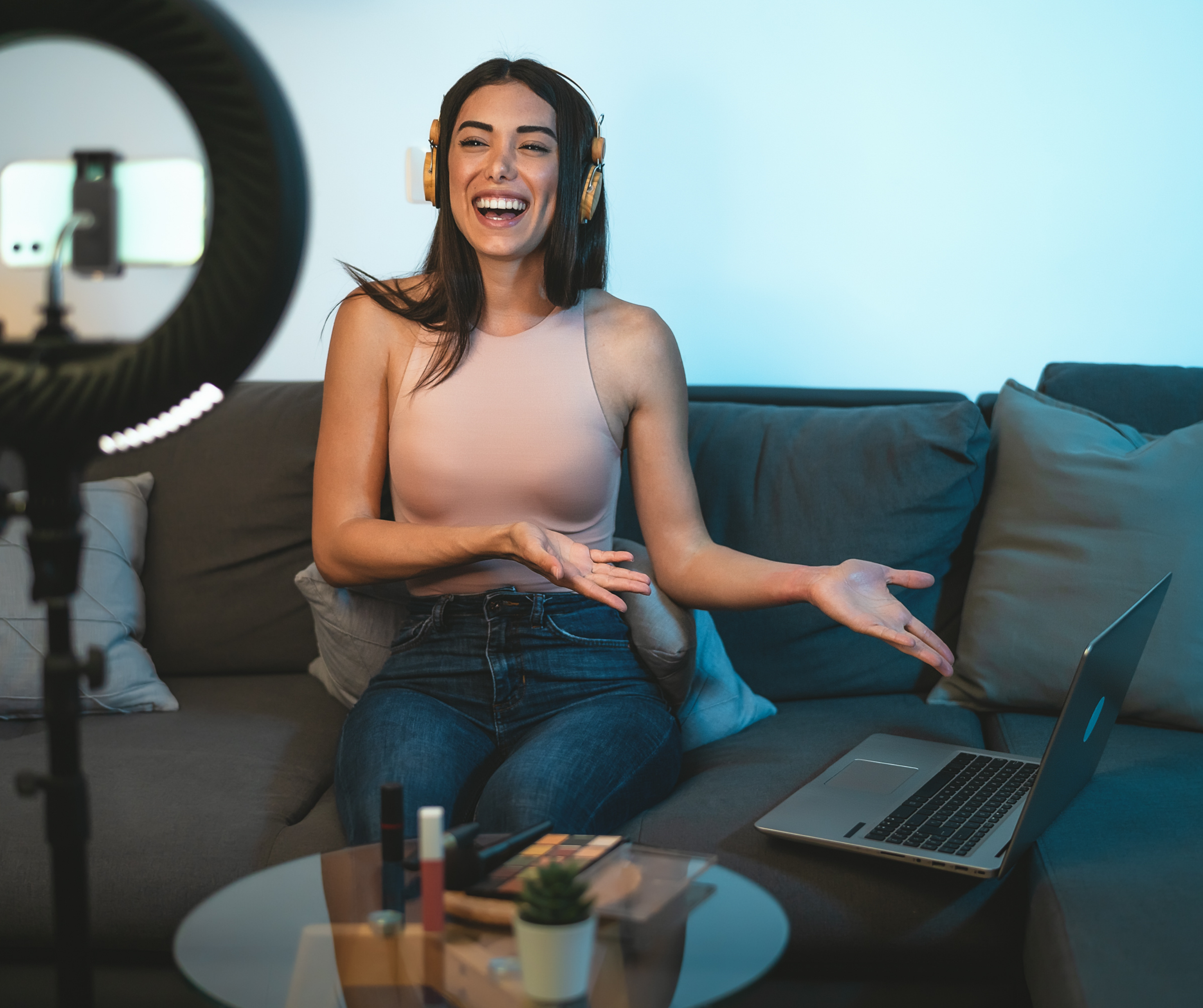 Woman wearing headphones sitting on a couch, engaging in a live stream with a ring light, smartphone, and laptop in view.