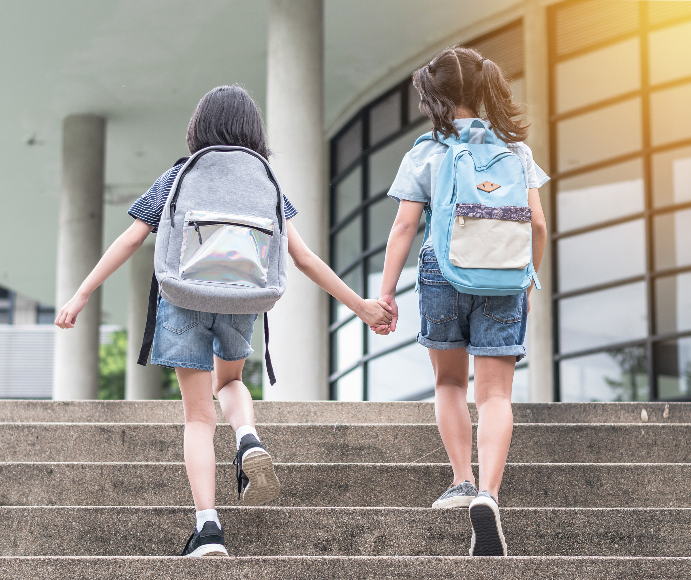 Two children with backpacks walking up the steps to a school building, holding hands.