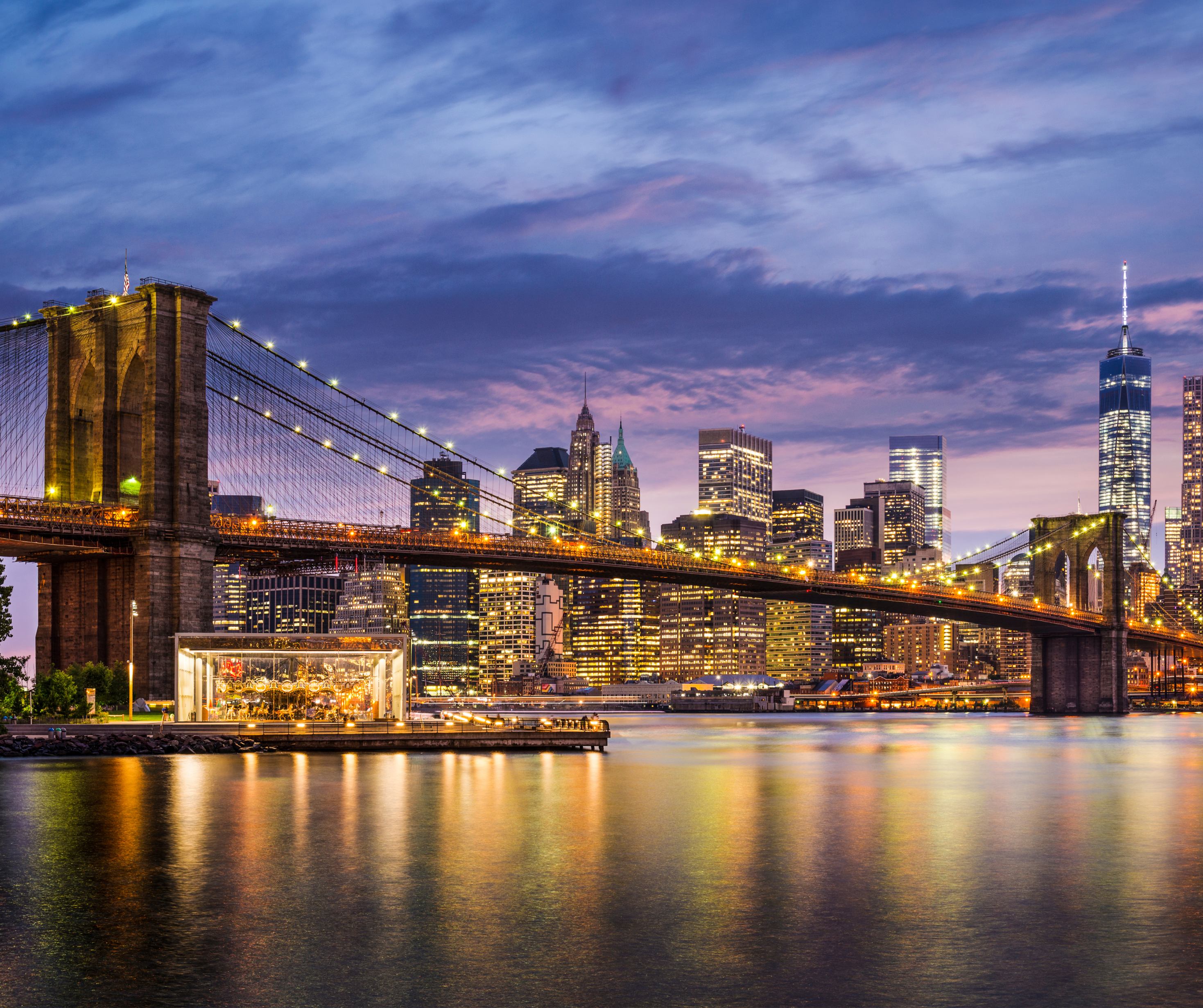 A scenic view of the Brooklyn Bridge leading into Manhattan, with the city skyline lit up at twilight.