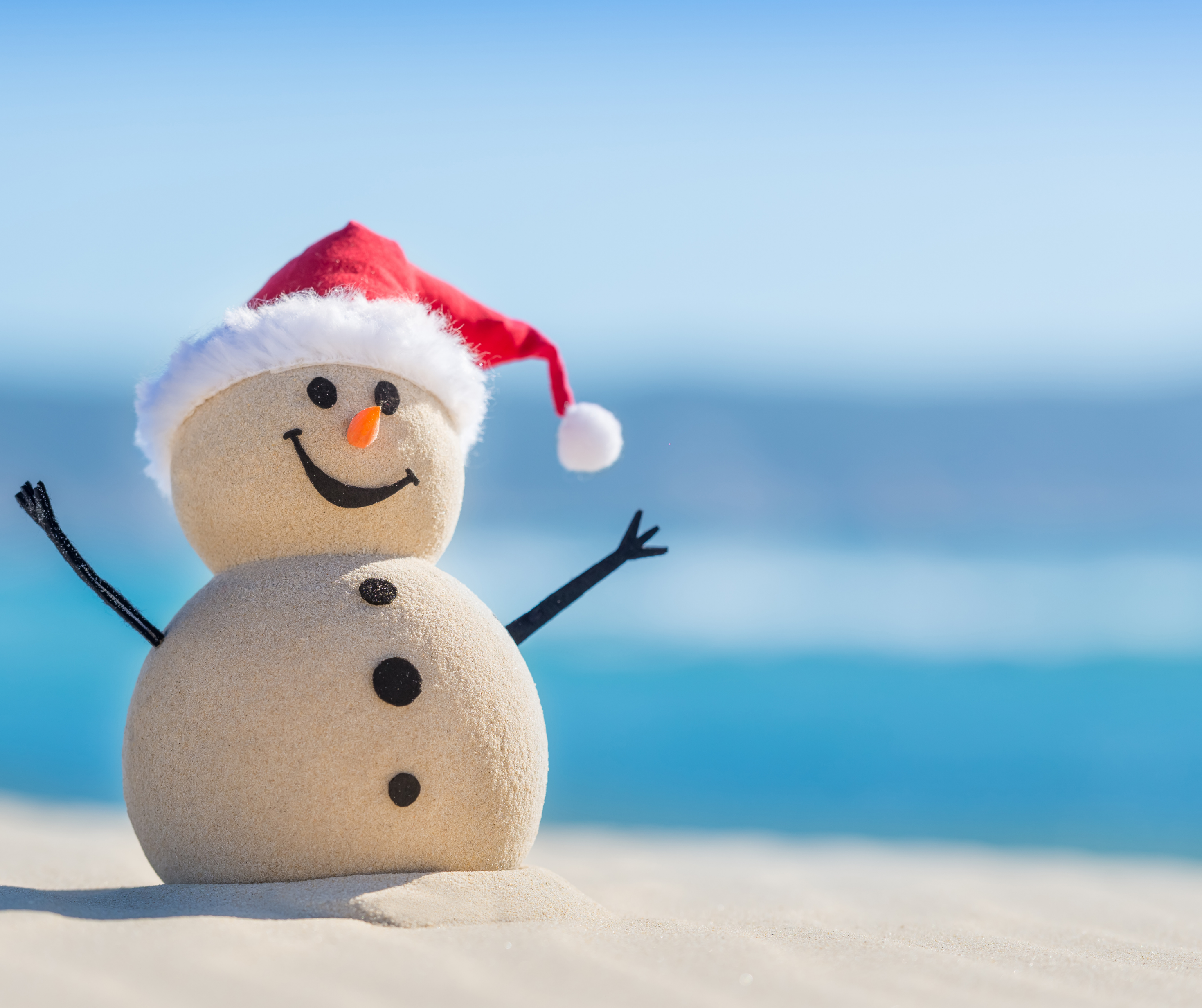 Sandman wearing a Santa hat on a beach with blue sea and sky in the background.