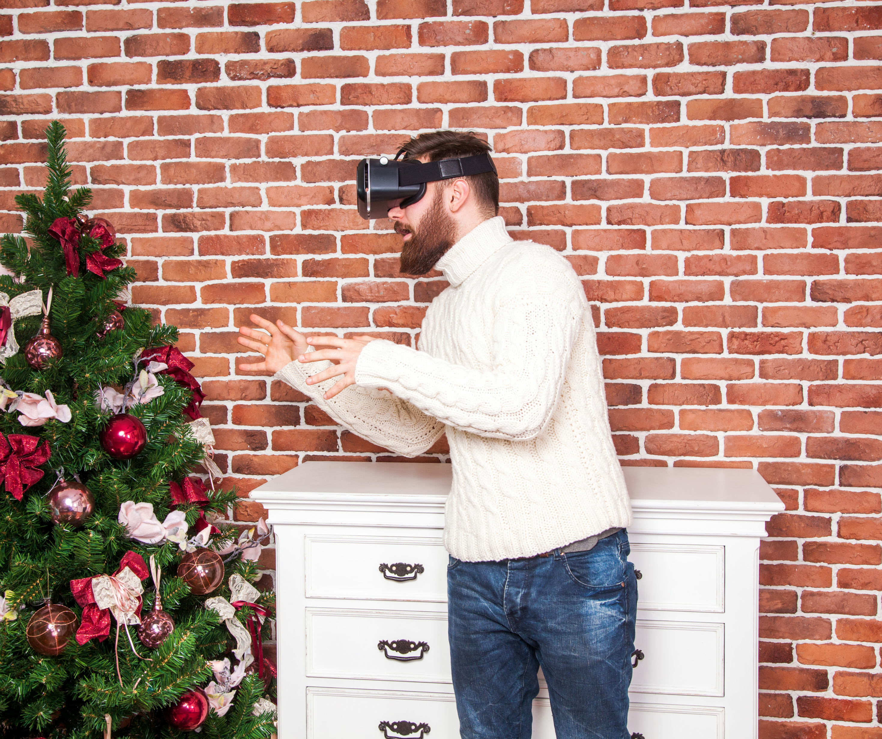 A bearded man in a white sweater interacts with a virtual reality headset near a decorated Christmas tree.