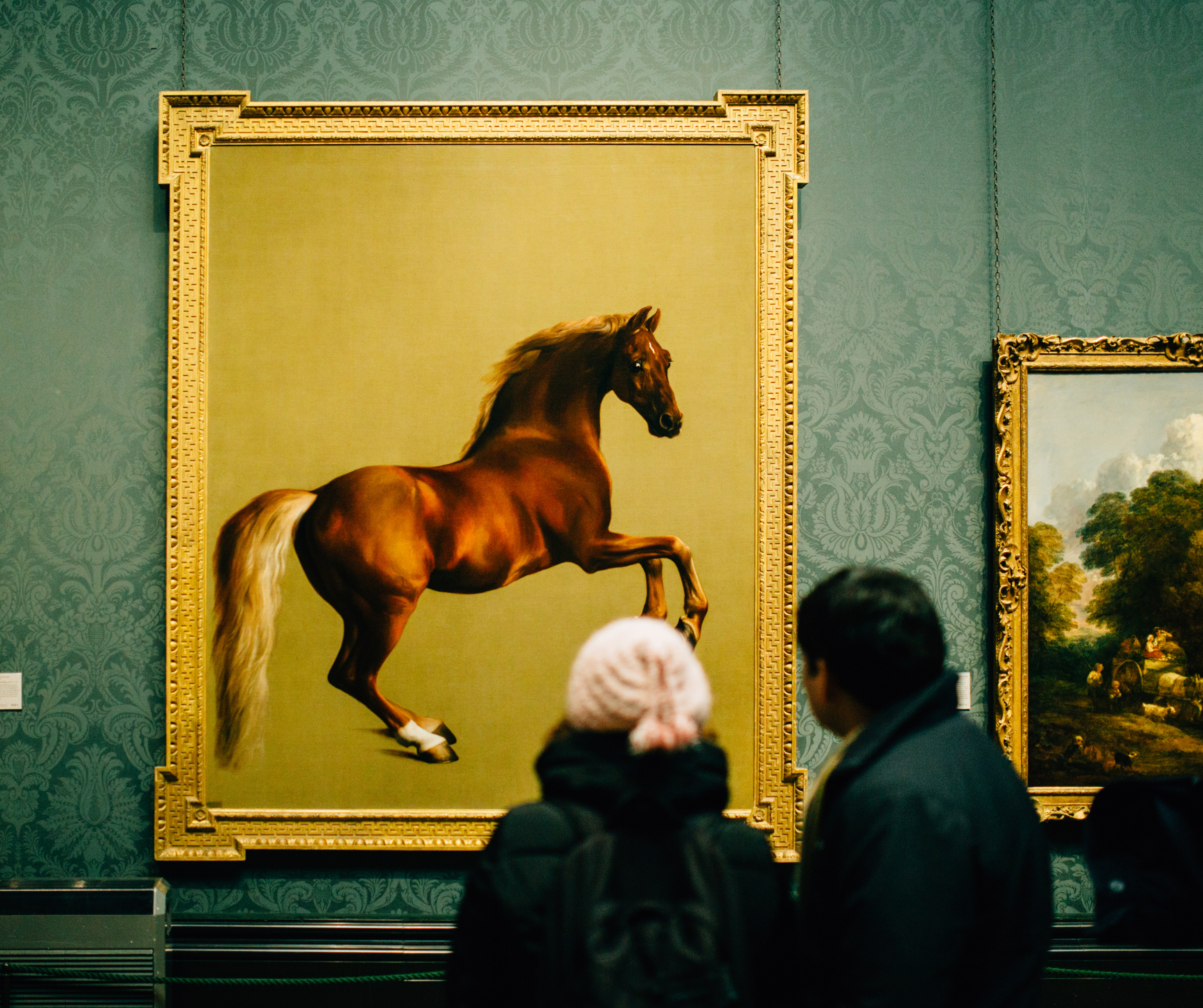 Two people viewing a large painting of a horse in an art gallery with ornate frames on the wall in Los Angeles