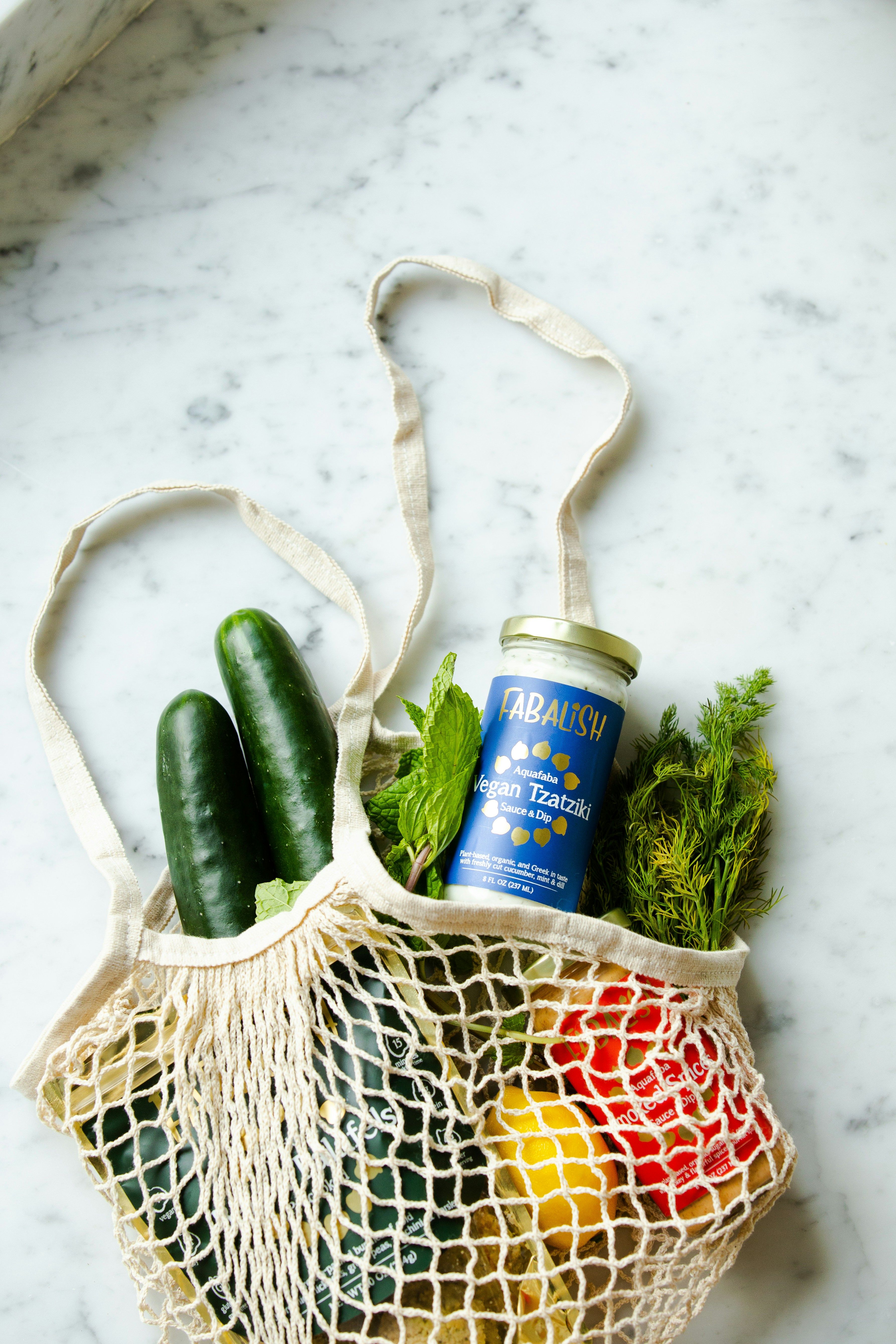 Cream-colored mesh shopping bag containing cucumbers, herbs, and a jar of vegan tzatziki