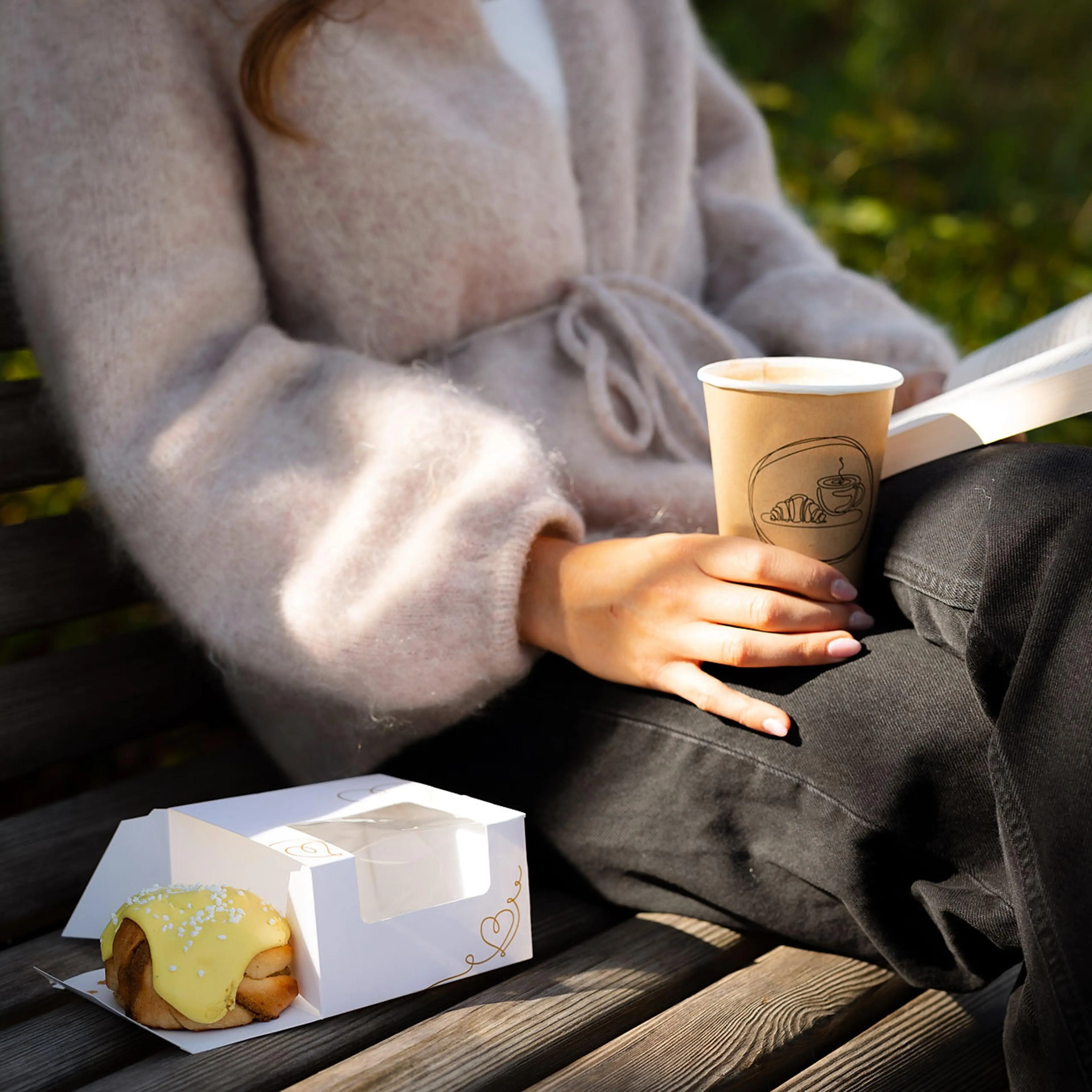 Kvinne leser bok på benk med Stund engangskopp og kakeeske