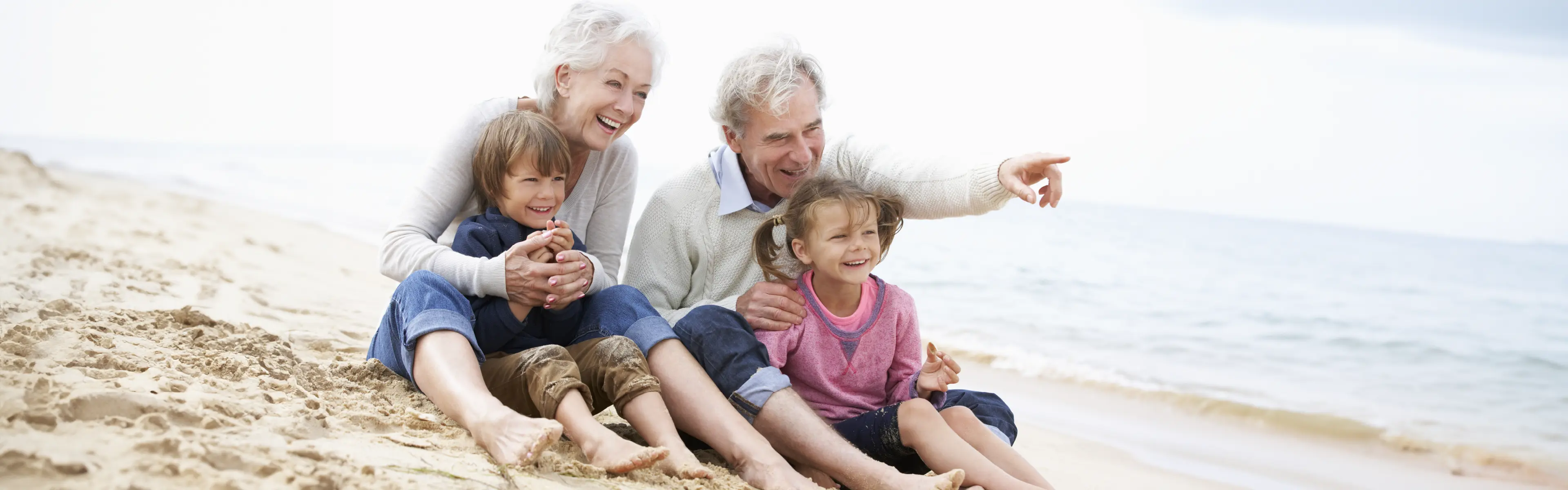 besteforeldre og barnebarn smilende på stranda, bestefar peker på noe utenfor bildet