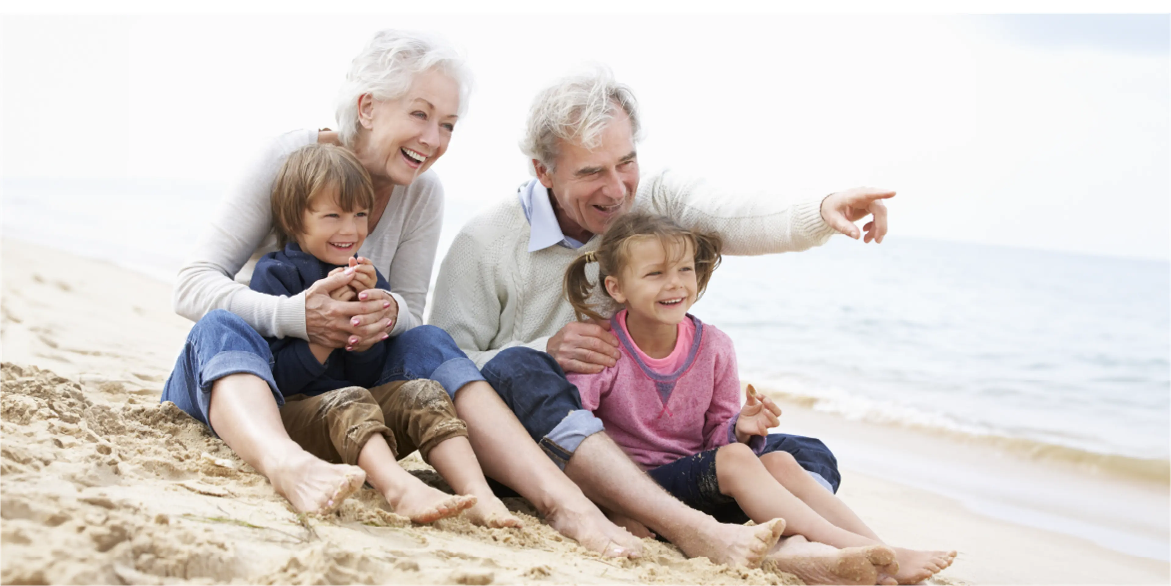 besteforeldre og barnebarn smilende på stranda, bestefar peker på noe utenfor bildet