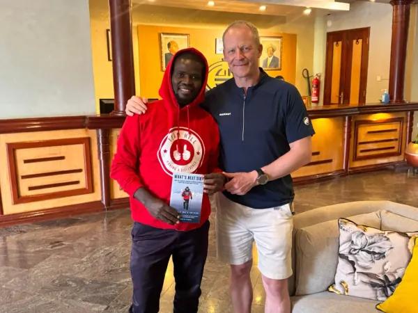 Two men posing indoors, one in a red hoodie holding a pamphlet