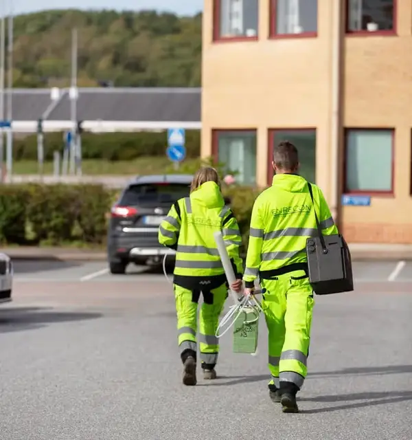 Två medarbetare i varselkläder går med provtagningsutrustning