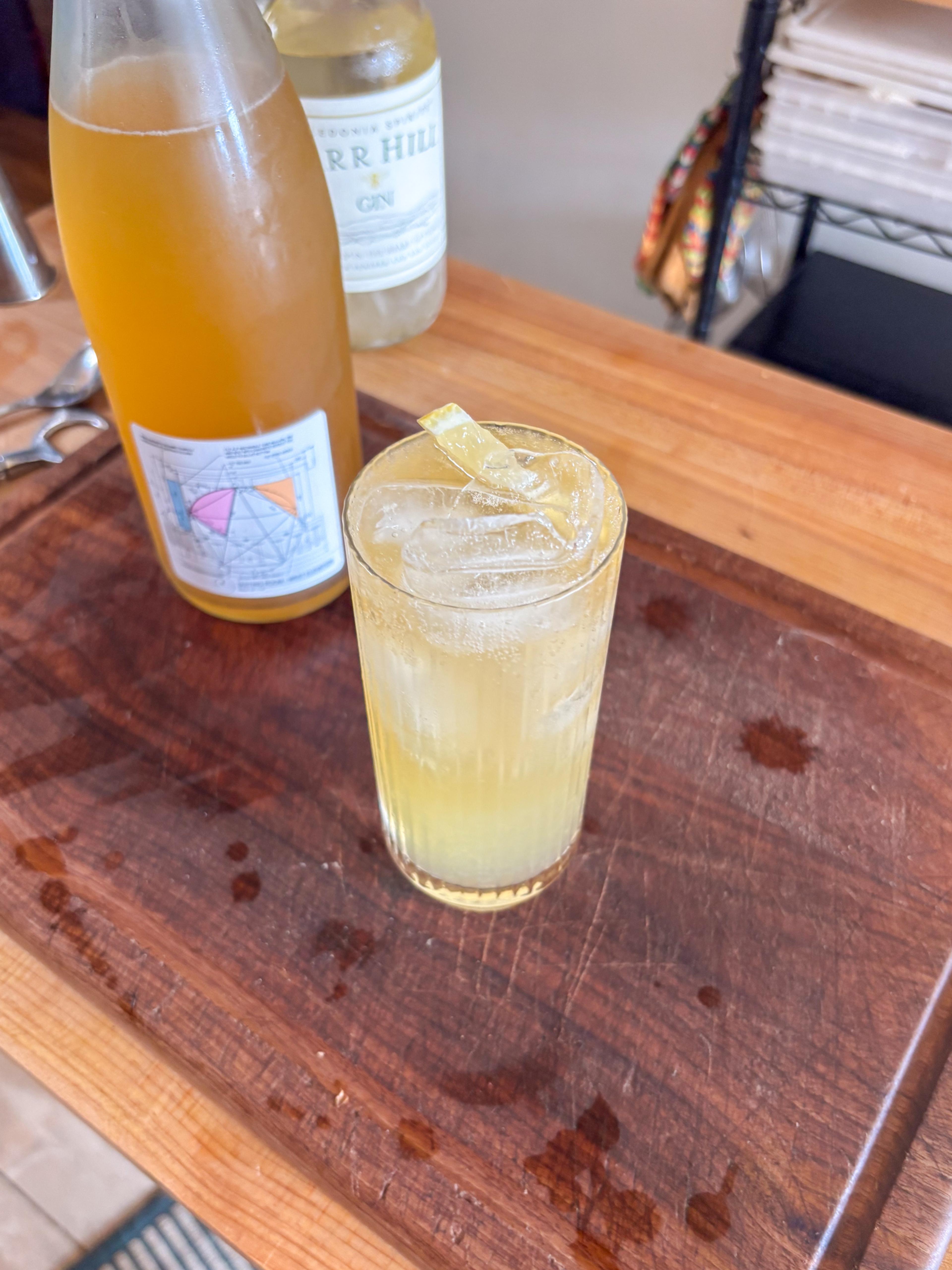 Picture of Mead Tom Collins cocktail in a collins glass, with a bottle of sparkling mead and gin behind it.