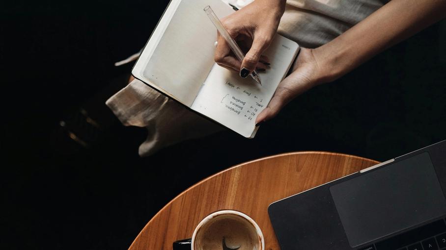 Woman writing in a notebook beside coffee and laptop, symbolizing reflection and planning before an oncology appointment.