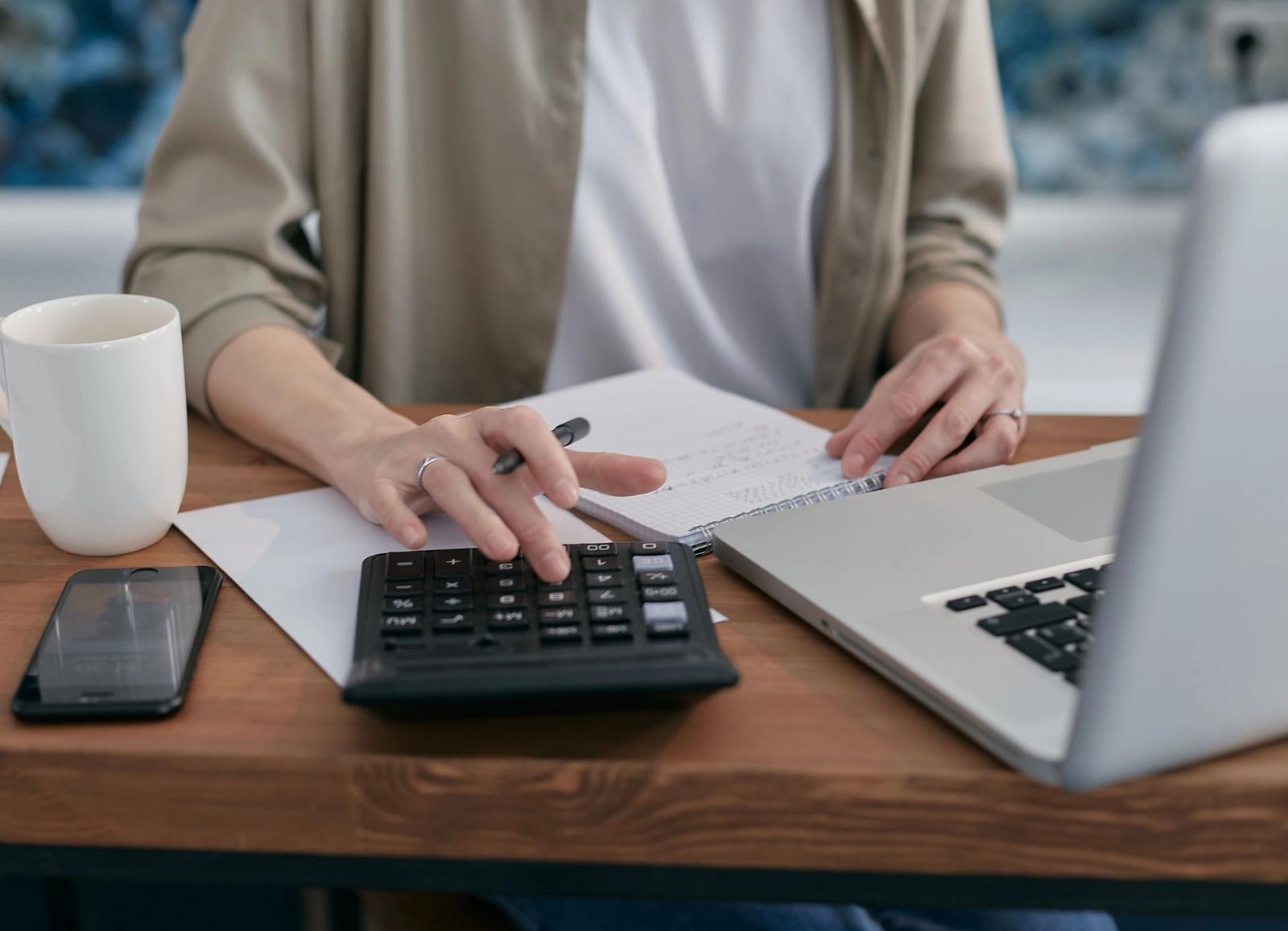 A person working at a laptop with a calculator, pen and notepad