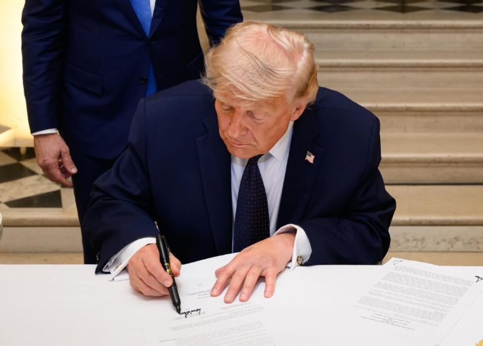Donald Trump is seated at a table and signing the memorandum of understanding in France