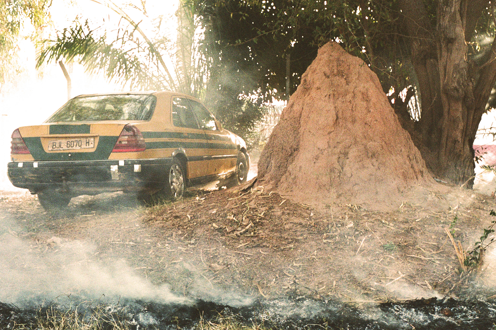 An outdoor, sun-drenched scene features a large, conical, reddish-brown termite mound in the foreground. To its left, a yellow sedan with a green horizontal stripe—resembling a West African taxi—is parked on a dusty, uneven dirt path. Smoke drifts across the bottom of the frame from a small patch of smoldering grass in the foreground. In the background, lush green trees and foliage are partially obscured by a bright, hazy light.