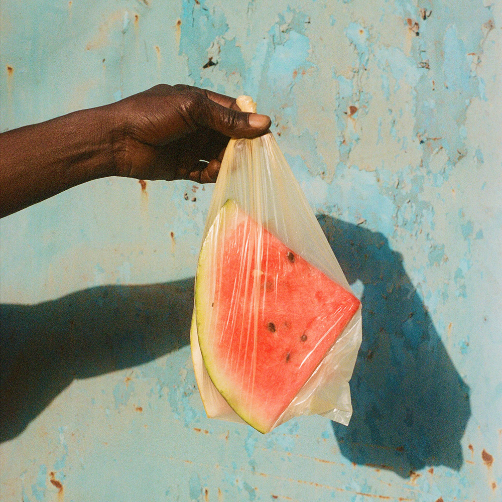An arm with dark skin extends from the left, its hand gripping the tied top of a clear plastic bag that contains a large, vibrant slice of watermelon.