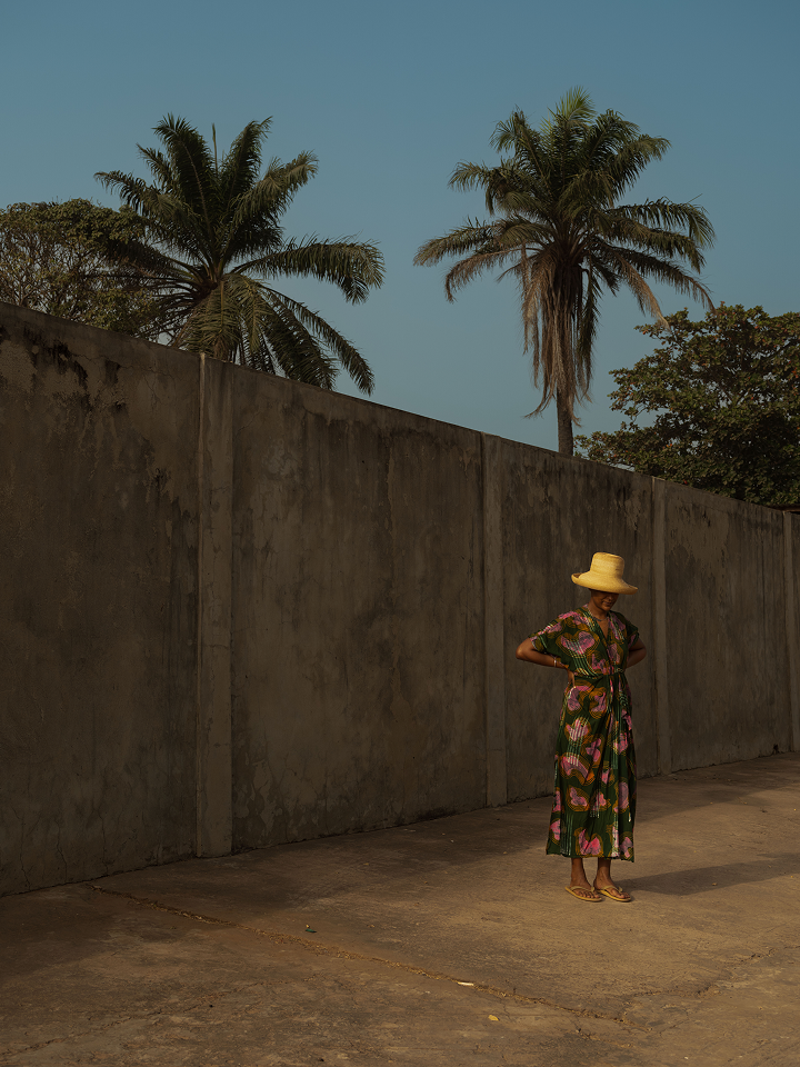 An individual stands in the shadow of a high, weathered concrete wall that stretches across the frame. They are wearing a long, green patterned dress with a tied waist and a wide-brimmed straw hat that casts their face in shadow. In the background, two tall palm trees and other lush greenery rise above the wall against a clear, pale blue sky. The scene is illuminated by warm, low-angled sunlight, creating long shadows on the ground.