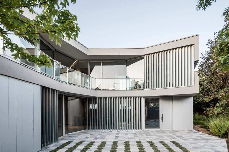 Exterior façade of a contemporary single-family home with architecture defined by organic lines and elegant volumetry that combines minimalism, functionality, and integration with the surroundings. The project stands out for its use of large glass surfaces, transparent railings, and vertical slats that provide privacy, solar control, and visual rhythm to the architectural composition. The paved outdoor spaces and perimeter vegetation reinforce the sense of spaciousness and connection with the landscape, while the light-toned finishes and continuous materials convey a modern, bright, and sophisticated aesthetic typical of a high-end architectural residence.