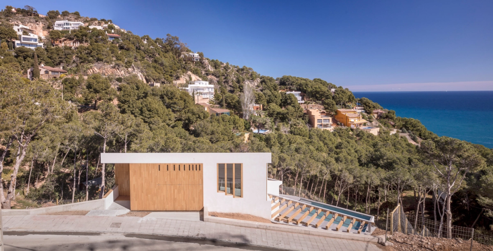 Detached single-family home located in a privileged setting with open sea views and integrated into the natural landscape of the Costa Brava. The project embraces minimalist and horizontal architecture, with pure volumes, white finishes, and wood cladding that provide warmth and elegance. The positioning on the site takes advantage of the sloping plot to create a fluid relationship between architecture, nature, and panoramic views. A contemporary detached house designed to enhance natural light, privacy, and a constant connection with the outdoors.