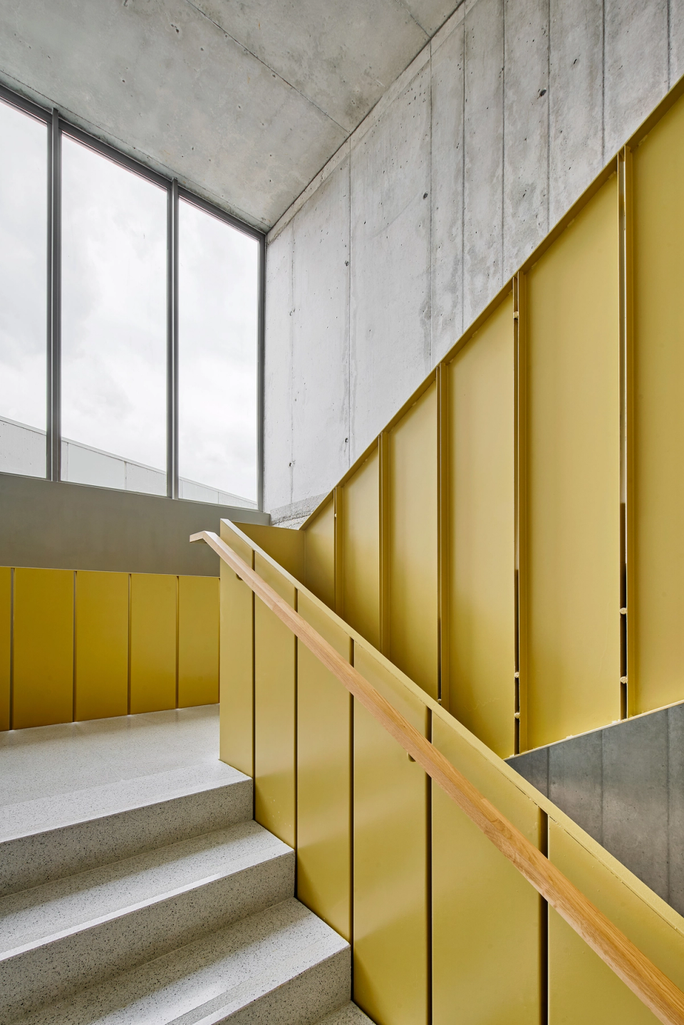 Interior staircase of a cultural center with contemporary architecture based on exposed concrete, natural light, and functional color details. The yellow-toned metal railings provide identity and dynamism to this vertical circulation space, while the large openings reinforce visual connection and the entry of light. This cultural center combines industrial materials and warm finishes to create a modern, accessible environment designed for community use.