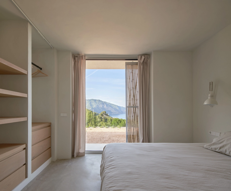 This bedroom in a single-family home embraces minimalist and functional architecture, with a clean layout that enhances natural light and views of the Mediterranean landscape. Vertical openings directly connect the interior space with the exterior, while cane filters and light curtains allow privacy and light entry to be naturally regulated. Neutral-toned finishes, integrated furniture, and the presence of light wood reinforce a serene and welcoming atmosphere. This single-family home stands out for its ability to integrate comfort, simplicity, and connection with the natural surroundings within contemporary and timeless architecture.
