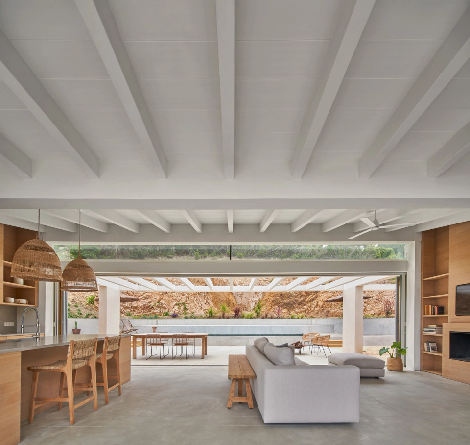 This single-family home embraces open and fluid architecture, where interior and exterior spaces connect naturally through large sliding enclosures. The daytime area integrates the kitchen, dining room, and living room into one spacious and bright environment, enhancing functionality and the visual relationship with the outdoor terrace. White-painted wooden beams, natural finishes, and simple-lined furniture bring warmth and reinforce the Mediterranean character of the home. This single-family home stands out for its natural light, spatial continuity, and contemporary architecture designed to enjoy the landscape and outdoor living.