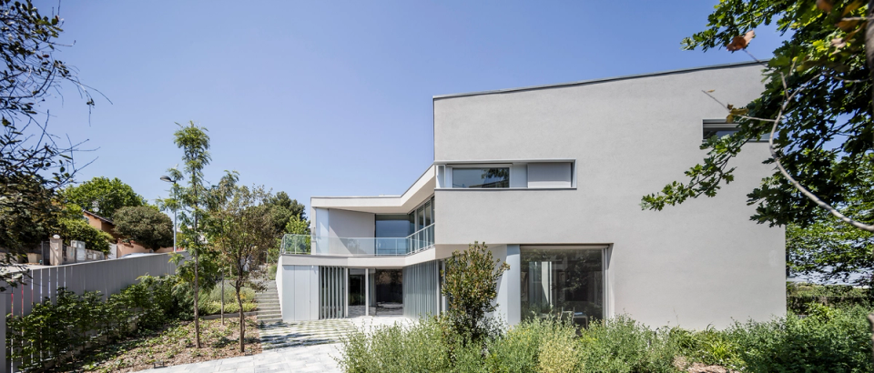 "Contemporary single-family house with an architectural composition of straight lines and refined volumes that integrates harmoniously with the natural surroundings. The façade, with light and minimalist finishes, combines large glazed openings, terraces, and vertical elements that provide depth, brightness, and privacy to the interior spaces. The exterior landscaping and perimeter vegetation reinforce the elegant and functional character of the home, enhancing the fluid relationship between architecture, landscape, and residential comfort.