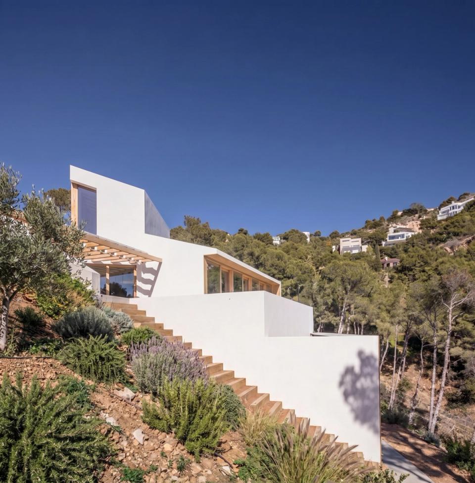Contemporary detached single-family home integrated into a sloping plot surrounded by native Mediterranean vegetation. The project stands out for its architecture of pure volumes and geometric lines that adapt naturally to the site’s topography. White façades are combined with wood cladding and large openings that provide warmth, natural light, and a constant connection with the landscape. This detached house has been designed to dialogue with its surroundings, enhancing views, privacy, and landscape integration through an elegant, sustainable, and timeless architectural proposal.