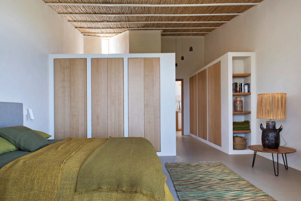 Bedroom of a detached house with large glass enclosures opening the space towards the terrace and outdoor landscape, enhancing natural light and the sense of calm. Ceilings clad with natural fibers, wooden carpentry and neutral tones create a warm, minimalist Mediterranean atmosphere fully integrated with the surrounding natural environment.