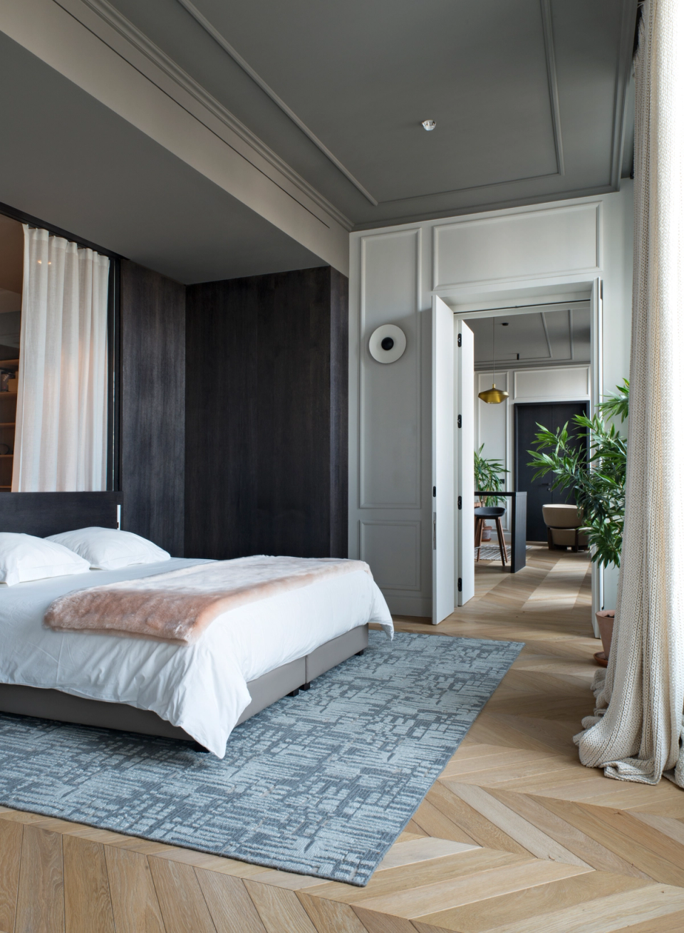 The interior renovation of this bedroom combines contemporary lines with classical architectural elements to create an elegant, balanced, and highly luminous space. The contrast between dark wood finishes and white molded walls adds visual depth and reinforces the sophisticated character of the home. Natural oak herringbone flooring, soft textiles, and natural light entering through the large windows create a warm and relaxed atmosphere designed to enhance comfort and well-being in the sleeping area.