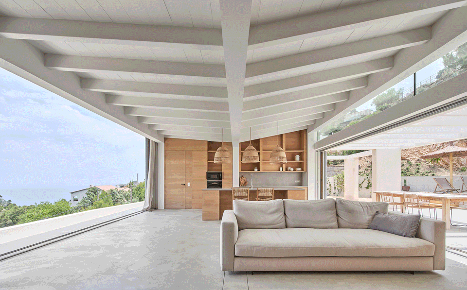 The interior of this single-family home has been conceived as a space completely open to the landscape, eliminating the boundaries between interior and exterior through large panoramic openings. The exposed white wooden structure brings rhythm and warmth to the spaces, while natural finishes and minimalist furniture reinforce a serene Mediterranean atmosphere. The living area and kitchen share one spacious and bright environment, designed to enhance the visual connection with the surroundings and the constant entry of natural light. This single-family home combines contemporary architecture, functionality, and landscape integration to create spacious, elegant, and comfortable spaces.