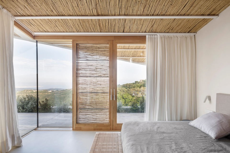Bedroom of a detached house with a serene and minimalist interior design that combines integrated natural wood furniture, neutral tones and warm materials to create a relaxing and welcoming atmosphere. Ceilings clad with natural fibers and the open spatial layout reinforce the sense of spaciousness, brightness and connection with the Mediterranean aesthetic of the home.