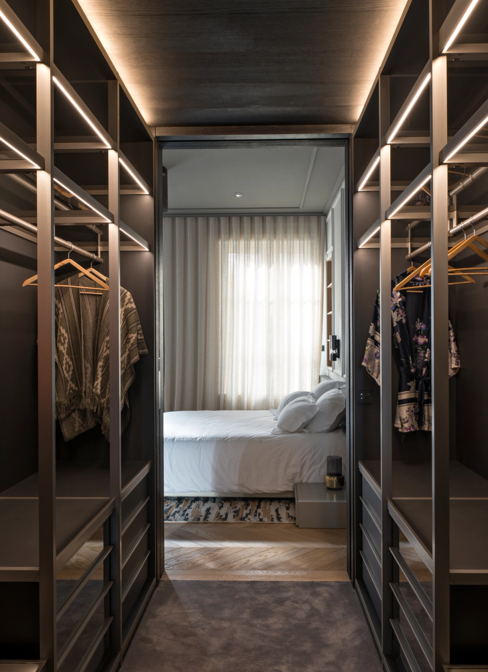 The interior renovation of this walk-in closet transforms the space into a functional and sophisticated area designed with a contemporary and elegant aesthetic. Dark finishes combined with integrated LED lighting on the shelves create an intimate and refined atmosphere that enhances the visual depth of the ensemble. The symmetrical layout and custom-made furniture optimize storage, while the visual connection with the bedroom adds continuity, spaciousness, and a sense of harmony throughout the home.