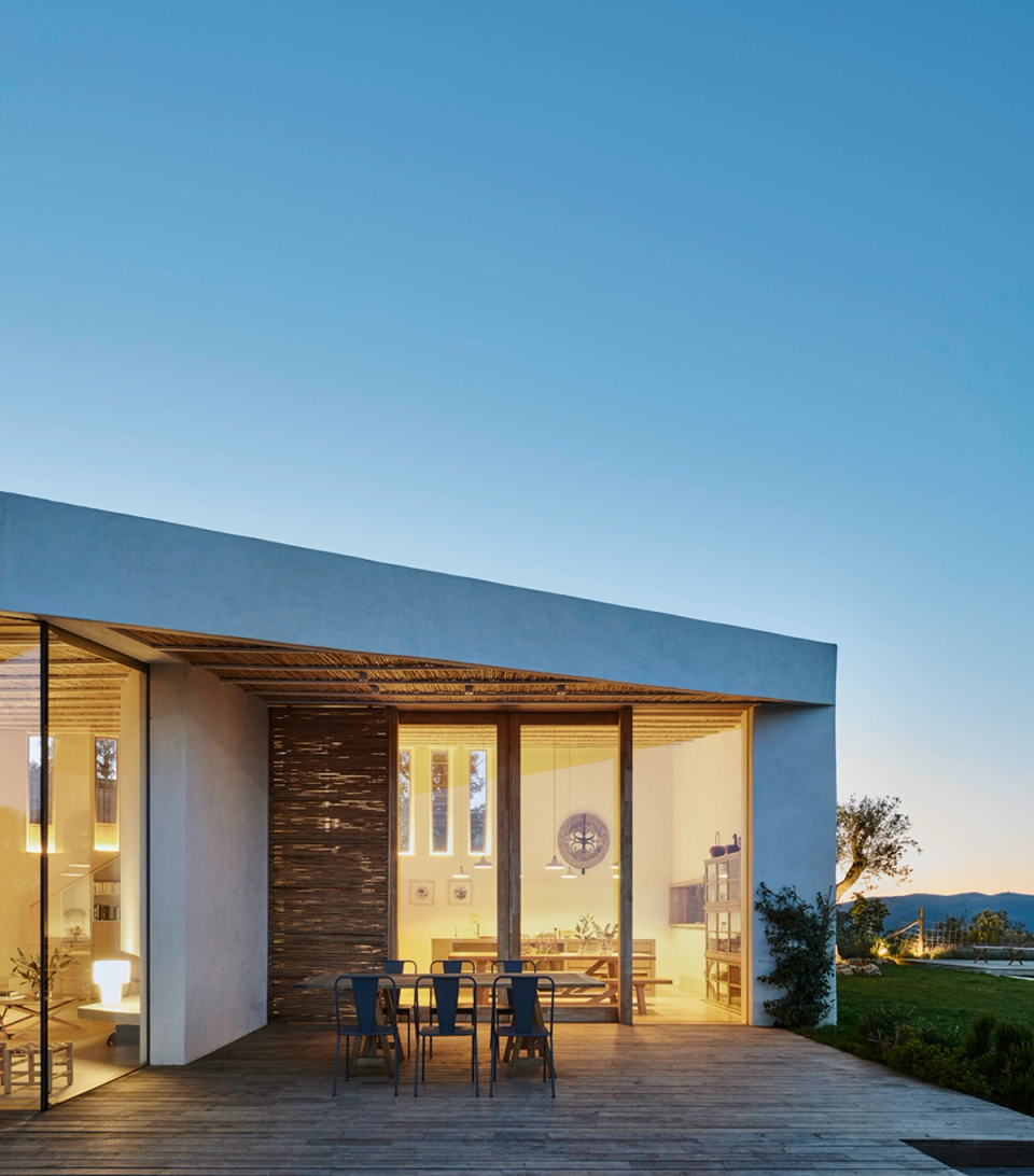 Exterior façade of a contemporary detached house with large glass openings and a wooden terrace integrated into the landscape. The warm interior lighting reinforces the connection between indoor and outdoor spaces, highlighting a minimalist architecture that combines clean lines, natural materials and a close relationship with the surroundings.