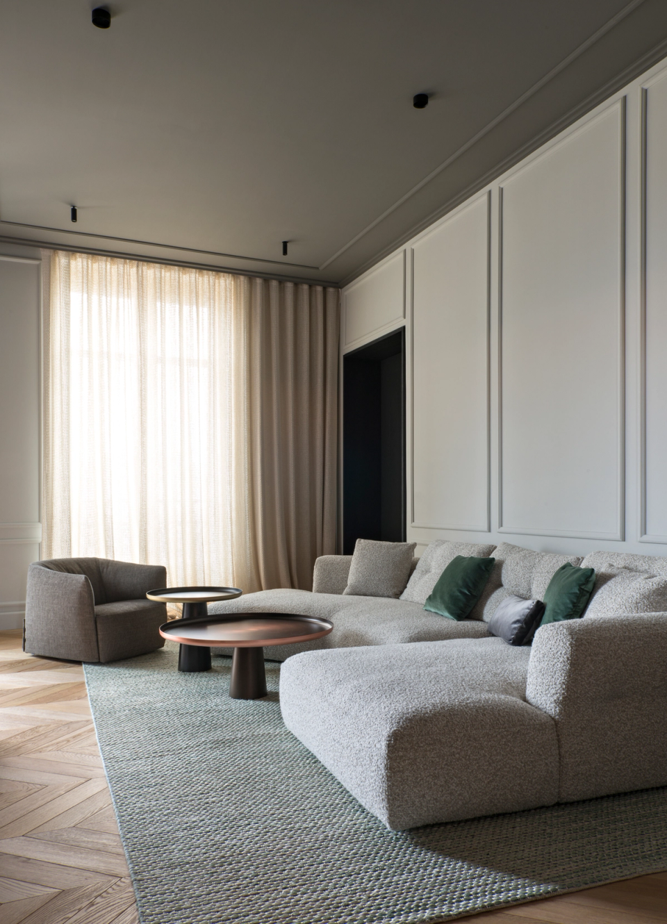 The interior renovation of this living room creates a serene and sophisticated atmosphere through a neutral palette, warm textures, and a carefully curated selection of contemporary furniture. Classical moldings and herringbone parquet flooring preserve the original essence of the home, while the soft volumes of the sofa, integrated lighting, and natural fabrics bring comfort and modernity. The light filtered through the curtains reinforces the sense of elegance and calm that defines the entire space.