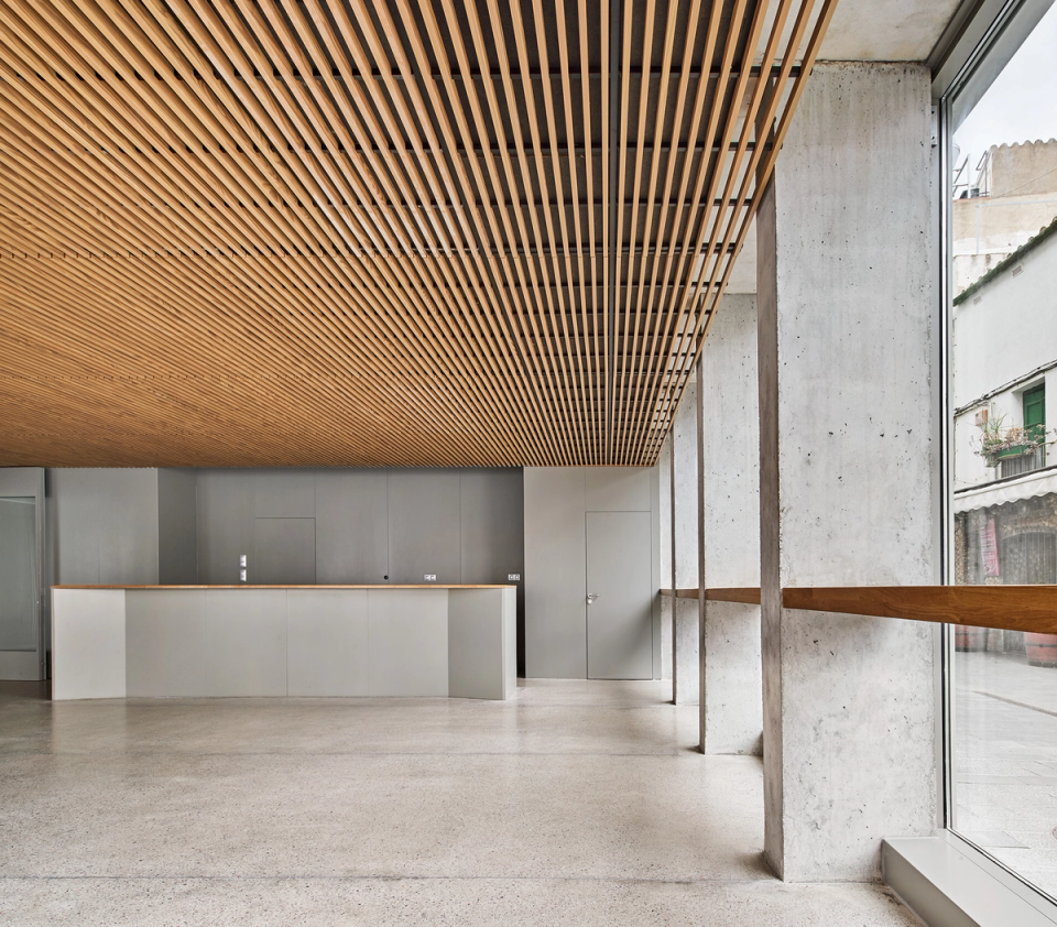 Interior space of a cultural center with a minimalist and contemporary aesthetic, highlighted by wooden slat ceilings and exposed concrete structural elements. Natural light and the open layout create a warm and functional atmosphere, ideal for reception, welcoming visitors, and hosting cultural activities. This cultural center combines noble materials and modern architectural solutions to create a versatile, elegant environment integrated with the urban space.