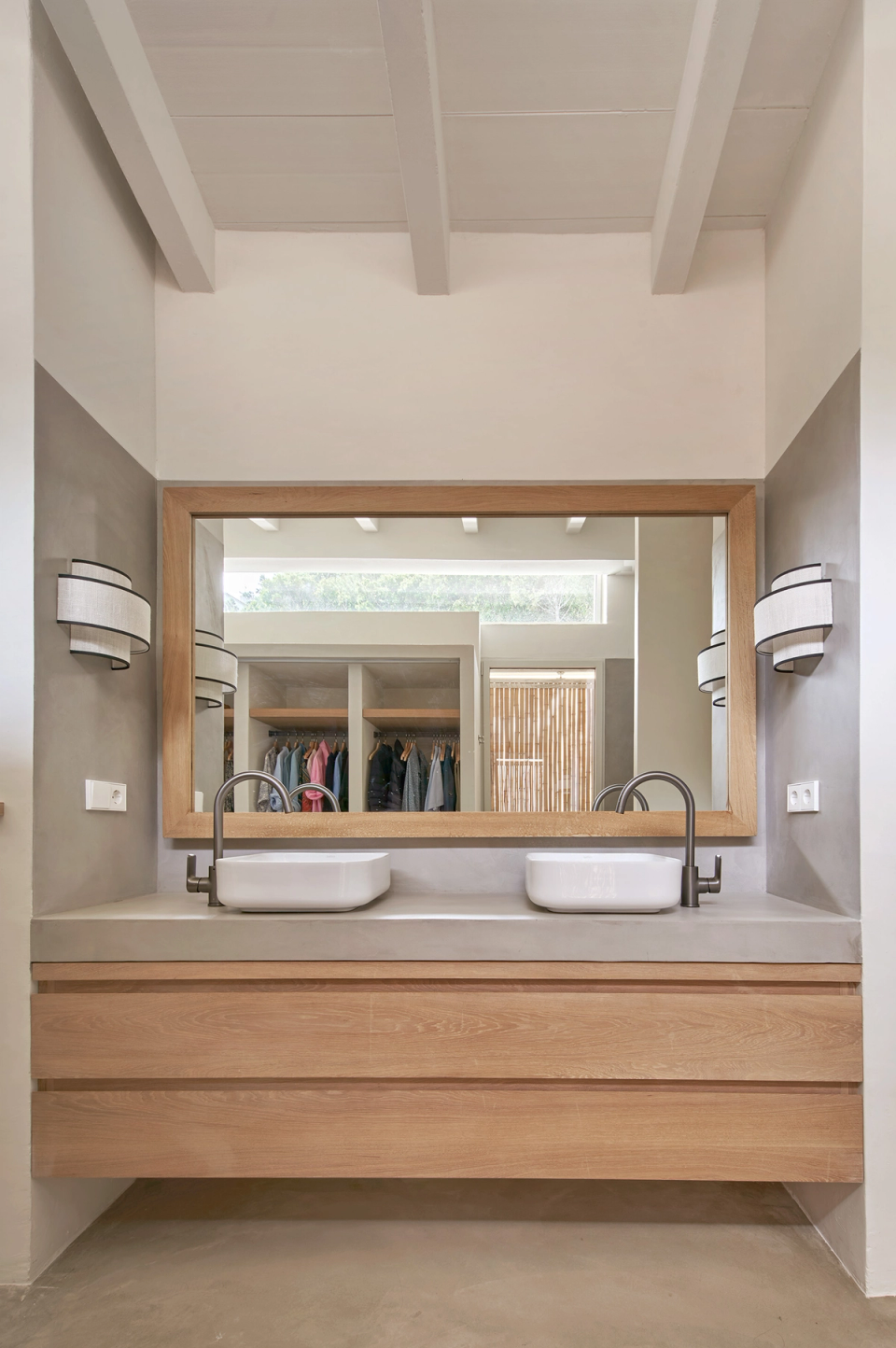 The bathroom of this single-family home reflects serene and functional architecture based on natural materials and a minimalist aesthetic. Suspended wooden furniture and continuous mineral-toned finishes provide warmth and elegance, while the large mirror frames the natural light and visually expands the space. Clean lines, dark-finish faucets, and integrated lighting reinforce the contemporary character of the project. This single-family home prioritizes everyday comfort and visual cohesion between all rooms, maintaining a constant relationship between architecture, interior design, and the Mediterranean landscape.