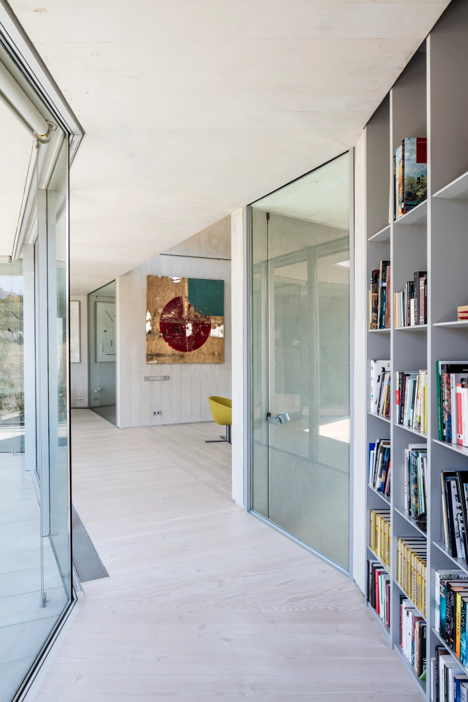 Corridor of a modern single-family home with a longitudinal layout that enhances natural light and transparency between spaces. Fully glazed façades allow a constant visual connection with the outdoors, while the combination of exposed concrete, light wood flooring, and glass enclosures creates a contemporary and elegant atmosphere. The integrated bookshelf along the circulation path provides functionality and character to a minimalist interior conceived to offer spaciousness, visual continuity, and architectural comfort.