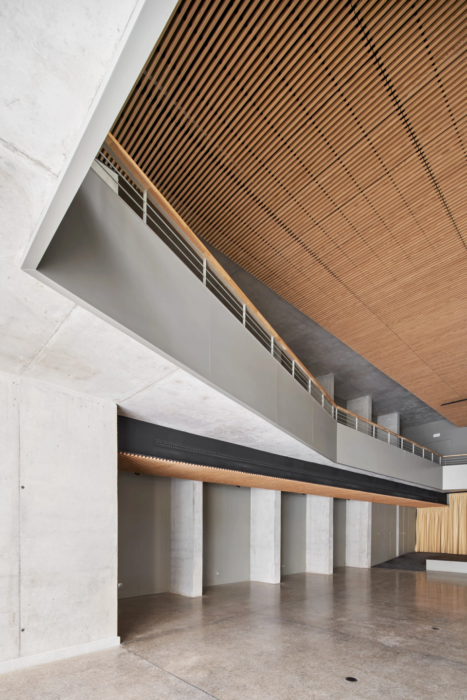 Interior view of a cultural center with contemporary architecture defined by clean lines, large cantilevers, and acoustic wood cladding. The combination of slatted technical ceilings, exposed concrete, and open spaces creates an elegant and functional atmosphere designed to host cultural activities, performances, and public events. The integrated stage and open layout reinforce the versatility of the facility, turning this cultural center into a flexible space adapted to multiple community uses.