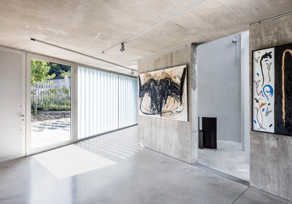 Interior of a contemporary single-family home featuring exposed concrete, open-plan spaces, and generous natural light. Large glazed openings and exterior vertical filters create plays of light and shadow that bring dynamism and depth to the space. The minimalist architecture combines with large-format artworks and continuous materials to generate a sophisticated, modern atmosphere with a strong visual identity.