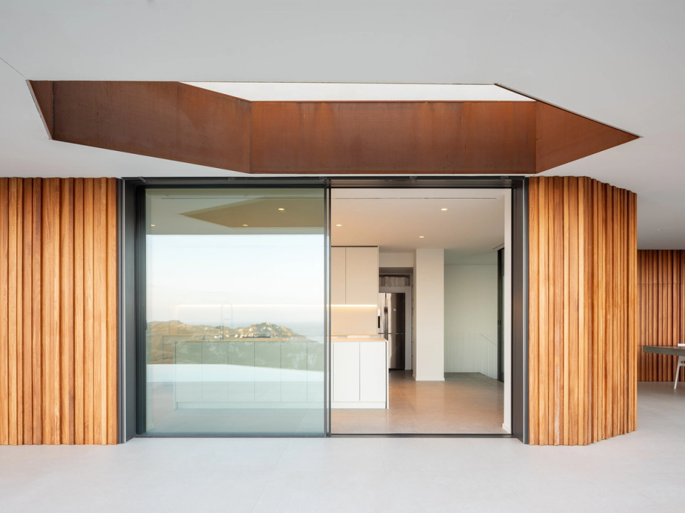 The detached single-family home features elegant and minimalist contemporary architecture defined by large white cantilevers, glass surfaces, and vertical wood cladding that adds texture and warmth. The covered outdoor area integrates completely with the interior through large sliding openings, connecting the kitchen and dining area with the terrace and infinity pool overlooking the landscape. The combination of natural materials, continuous flooring, and understated furniture creates a bright, refined atmosphere deeply connected to the Mediterranean surroundings.