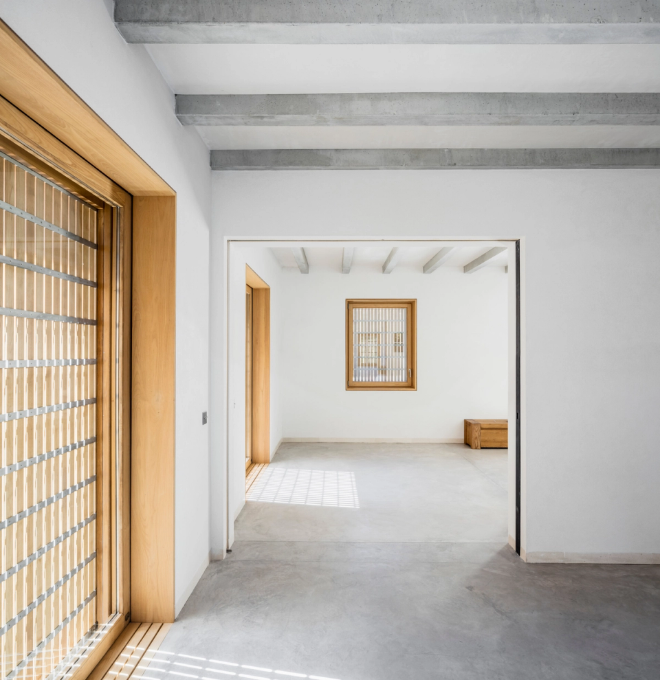 Interior spaces of a comprehensive renovation with minimalist architecture based on natural light, noble materials, and pure lines. Wooden-framed openings, exposed concrete beam ceilings, and continuous flooring create a serene, warm, and contemporary atmosphere. The open layout and visual connection between rooms enhance spaciousness and comfort, while solar protection elements provide privacy and a play of shadows that reinforces the architectural character of the home.
