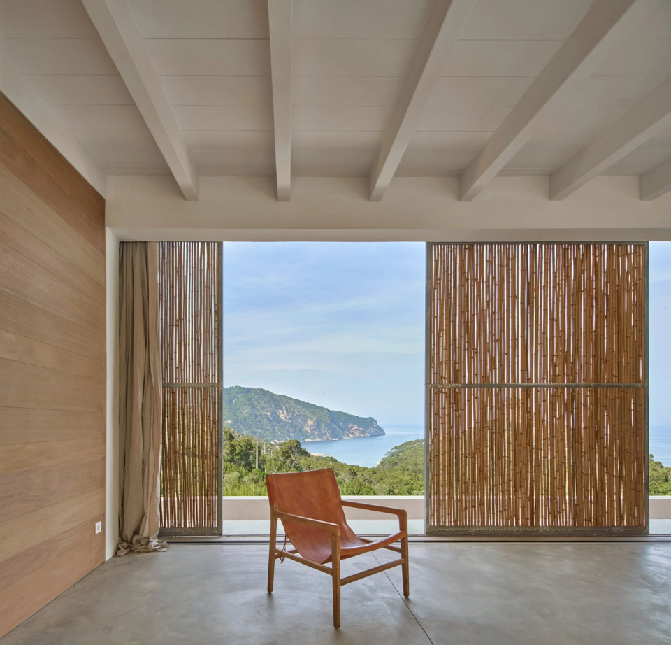 The interior of this single-family home stands out for its direct connection with the landscape and for an open architecture that prioritizes natural light and panoramic sea views. Large openings with sliding enclosures and cane filters allow the modulation of light entry and the creation of different atmospheres throughout the day, providing privacy and climatic comfort. Ceilings with exposed beams and natural wood finishes reinforce the feeling of warmth and continuity between interior and exterior. This single-family home combines minimalism, natural materials, and a constant relationship with the Mediterranean surroundings to create spacious, serene spaces fully integrated into the landscape.