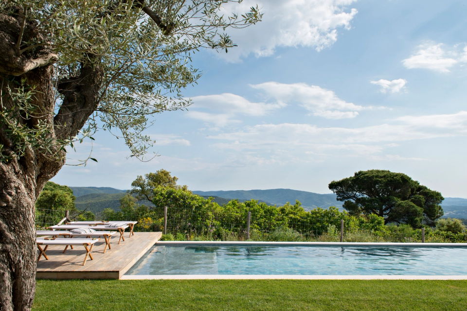 Exterior view of a modern detached house with an infinity pool and clean-lined architecture integrated into the landscape. The home stands out for its minimalist volumes, large glass surfaces and the combination of white tones and natural materials, creating continuous connections between interior spaces, the terrace and the Mediterranean environment.