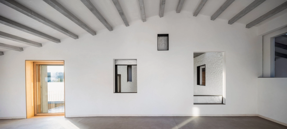 Interior space of a comprehensive renovation with minimalist lines, sloped ceilings with exposed concrete beams, and a layout designed to enhance natural light and visual connections between environments. Framed openings create architectural perspectives that provide depth and character, while the combination of white walls, continuous flooring, and natural wood carpentry generates a serene, contemporary, and balanced atmosphere. A full renovation that merges traditional materials and modern language with great spatial sensitivity.
