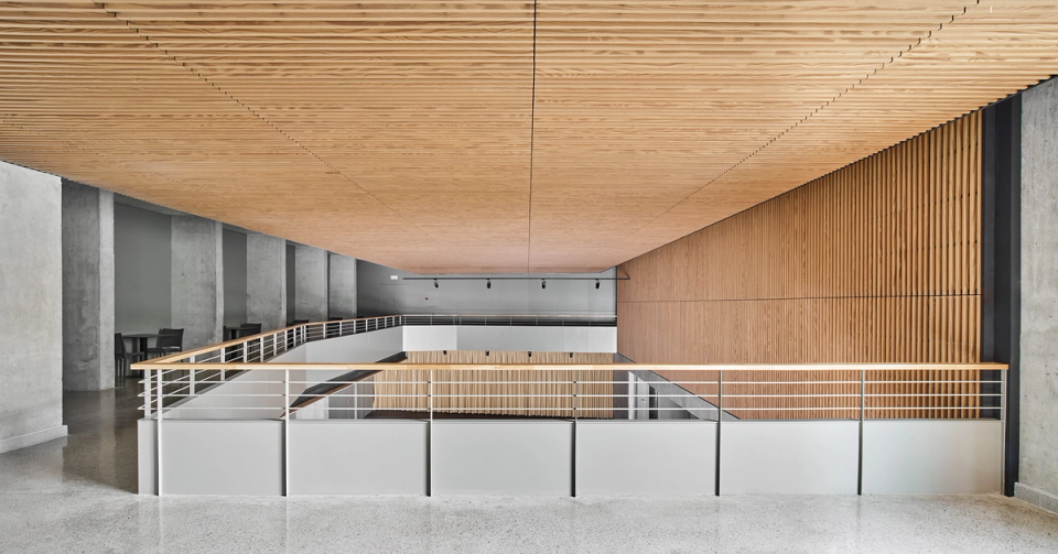 Upper space of a contemporary cultural center with interior architecture defined by wood cladding, horizontal lines, and a sense of visual spaciousness. The large acoustic slatted ceiling and minimalist metal railings create a modern and functional environment designed to facilitate circulation and connection between the different spaces of the cultural center. The combination of noble materials, natural light, and sober finishes provides elegance and versatility to this multipurpose cultural facility.