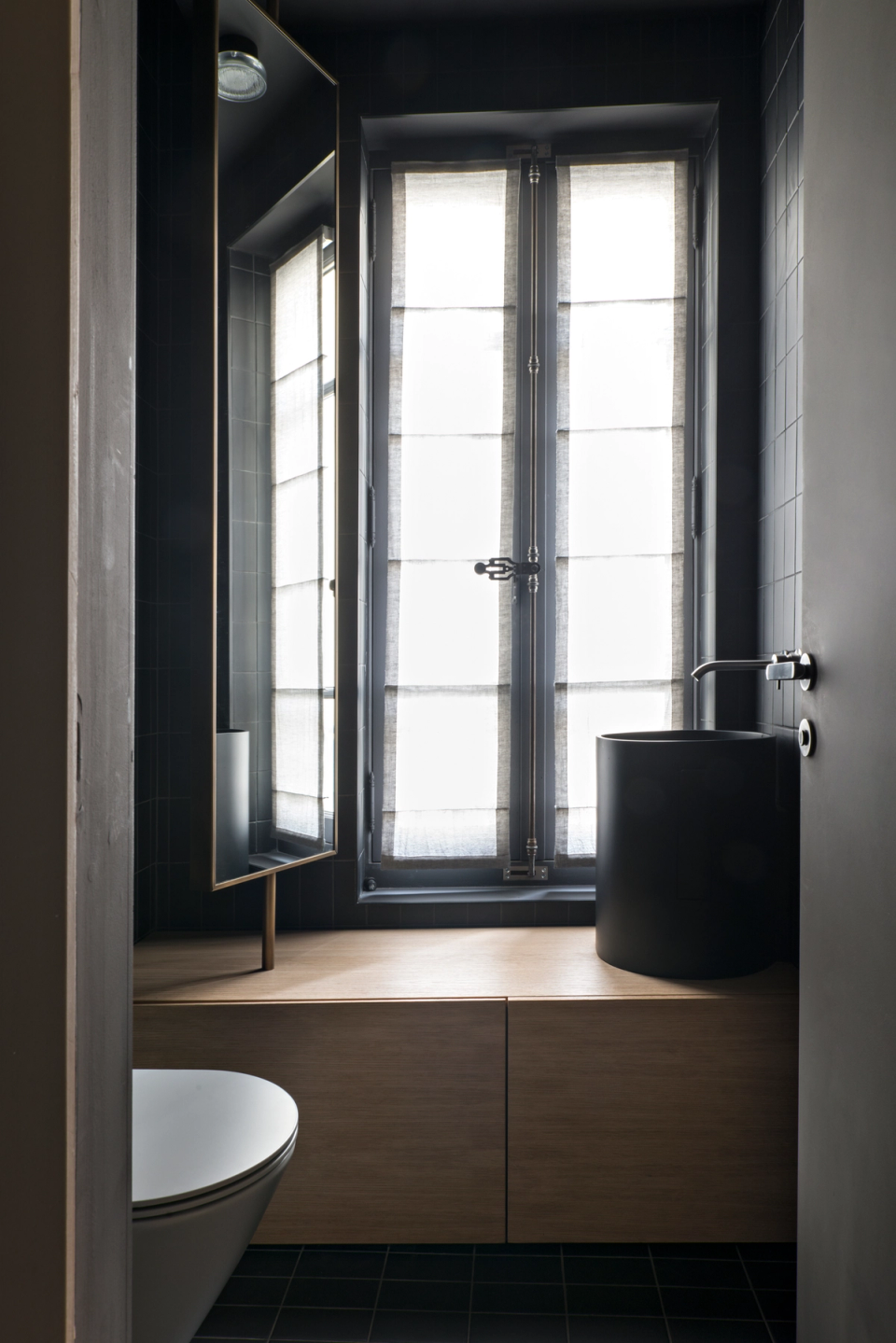 The interior renovation of this bathroom combines noble materials and a minimalist composition to create an intimate and sophisticated space. Natural wood adds warmth and contrasts with the dark surfaces, while the cylindrical sink and clean furniture lines reinforce the contemporary character of the design. Natural light filtered through textile blinds creates a soft and elegant atmosphere, enhancing the depth of the space and highlighting the attention to detail and textures.