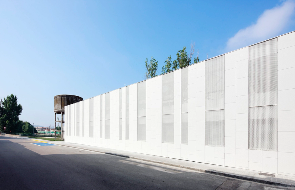 Side façade of the Sant Vicenç Health Center resolved through a minimalist and orderly composition, marked by white surfaces and vertical bands that provide rhythm, texture, and natural light filtering. The presence of the historic water tower in the background establishes a subtle contrast between industrial heritage and contemporary architecture, reinforcing the unique identity of the facility within the urban environment.