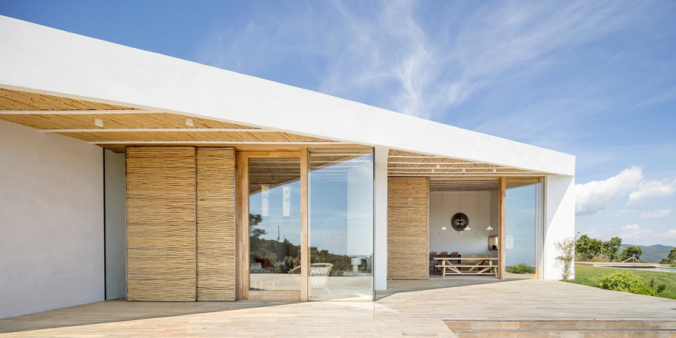 Exterior façade of a detached house with contemporary architecture open to the landscape, defined by large glass enclosures, spacious porches and natural wood cladding that provide texture and warmth. The pitched roof and outdoor spaces connect fluidly with the interior, enhancing natural light, views and integration with the Mediterranean environment.