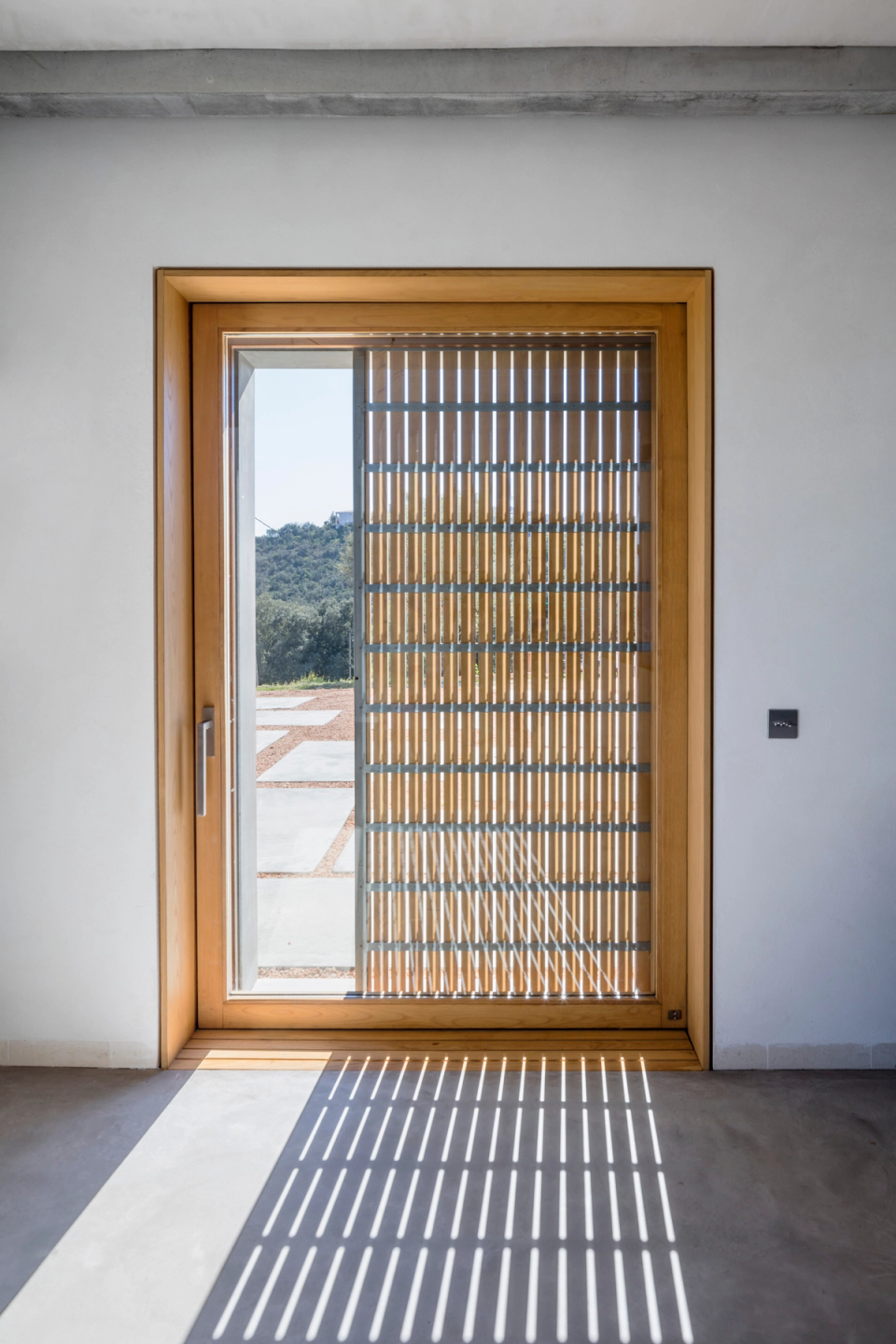 Architectural detail of a comprehensive renovation featuring natural wood enclosures and solar protection solutions that enhance the entry of natural light. The combination of minimalist lines, noble materials, and projected shadows creates an elegant, warm, and contemporary space. This full renovation stands out for the attention to finishes, the efficiency of the construction elements, and the visual connection between interior and exterior, providing comfort, brightness, and a timeless aesthetic.